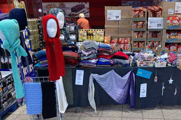 Ein Verkaufsstand mit handgefertigten Wollwaren, darunter Schals, Mützen und Tücher, liebevoll dekoriert. Der Stand, der sich in einem Supermarkt befindet, wird vom Kreativkreis des Hospizes betreut. Im Hintergrund sind weitere Produkte des Supermarkts zu sehen. Der gesamte Erlös der verkauften Artikel kommt dem Hospiz zum hl. Franziskus zugute.