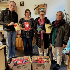 Vier Personen, die stolz die liebevoll gestalteten Geschenktüten in den Händen halten. Vor ihnen stehen Kartons voller weiterer Geschenke, die für eine weihnachtliche Aktion vorbereitet wurden. Die Gruppe lächelt in die Kamera.